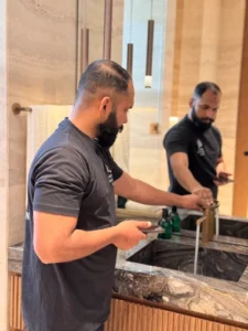 2026-04-21 2 - Property Inspection Company - Property Inspection Company Bearded man in a dark T-shirt turns on a bathroom sink faucet while looking at his phone in a modern bathroom.